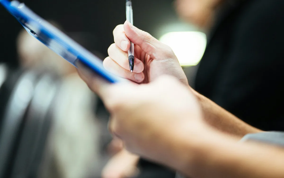 A hand holding a pen over a clipboard of printed checks